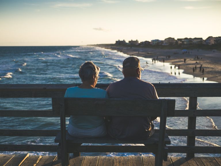 Back of an older couple on a bench facing a busy beach at sunset