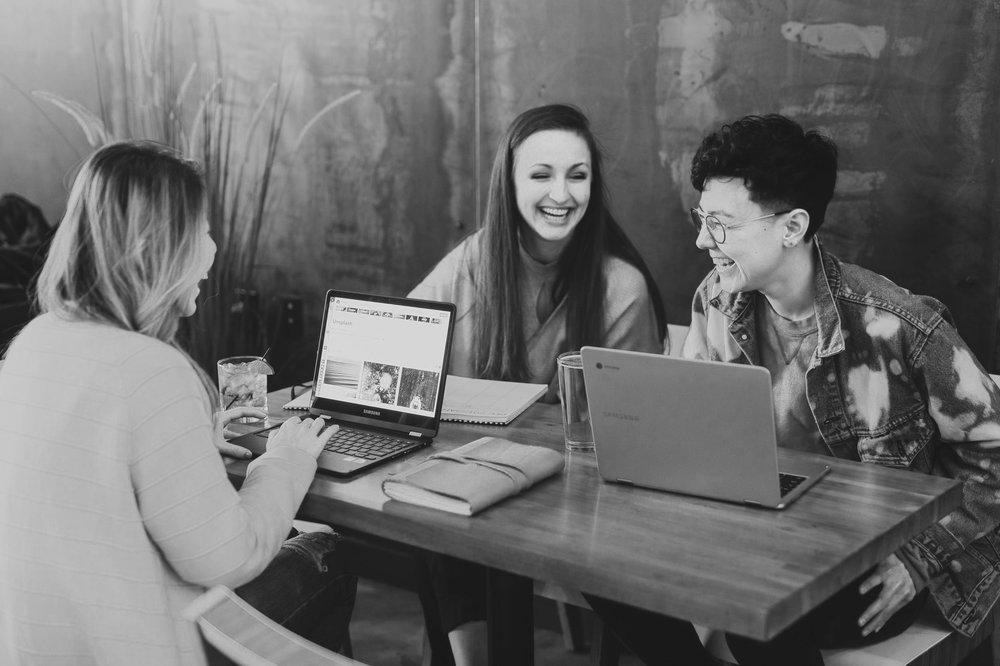 Black and white of three people at a coffee shop with laptops laughing